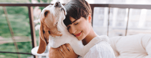 Woman smiling, hugging a Beagle, eyes closed in contentment, on a balcony with a blurred green cityscape in the background.