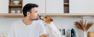 Man in white shirt kissing small dog in a kitchen, with shelves and dried plants in the background.