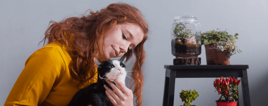 Woman with red hair cuddling a black and white cat next to a small table with potted plants and a jar terrarium.