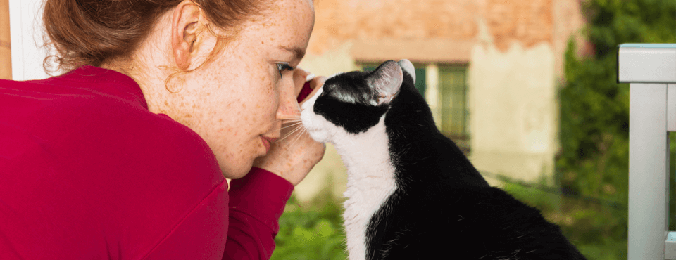 A woman in a red sweater touching noses with a black and white cat on a balcony with a blurred building in the background.