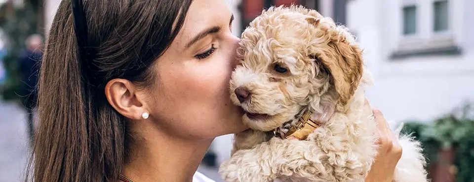 Woman with a ponytail lovingly kissing a fluffy, light brown puppy she's holding, in an outdoor setting.