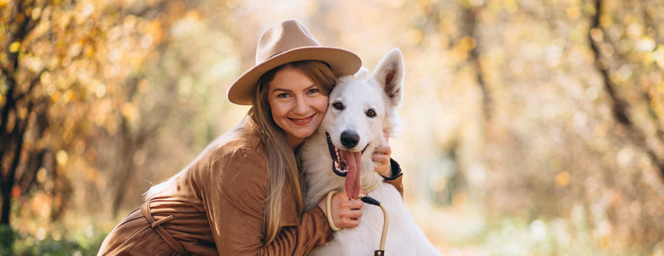 Woman in a brown hat and jacket smiling with a white dog in an autumn forest setting.